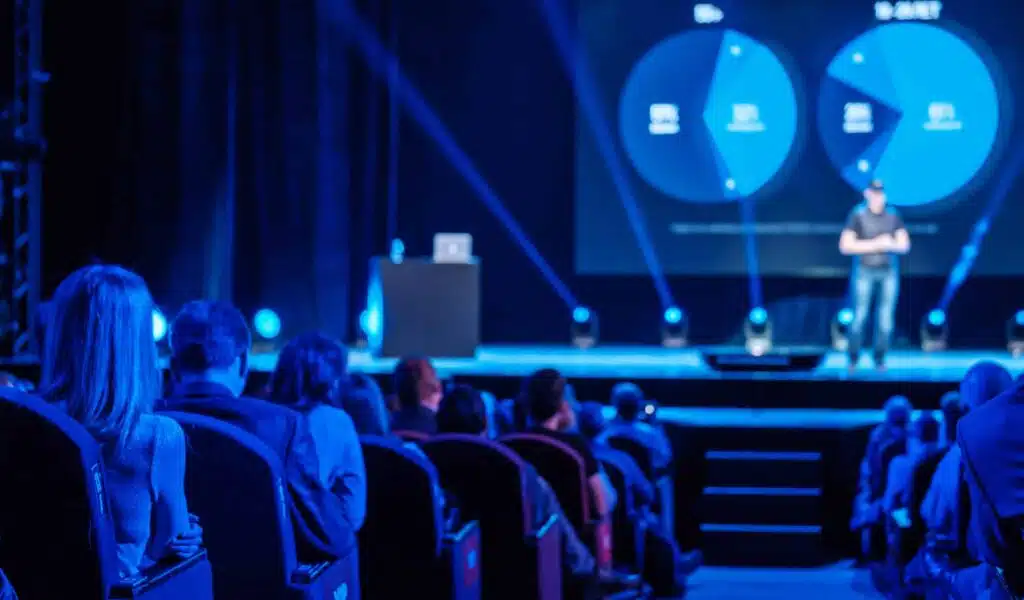 Pubblico che ascolta il relatore di una conferenza in una sala congressi illuminata in maniera scenografica (Allestimenti scenografici)